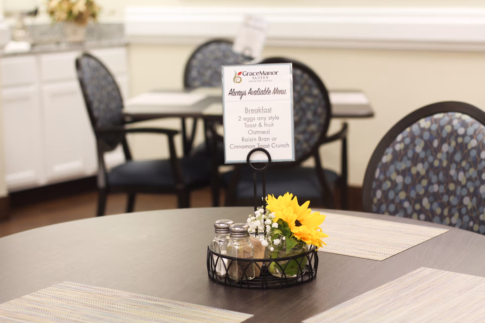 Round dining table with a centerpiece holding salt and pepper shakers, a small sunflower arrangement and a Grace Manor Suites menu card, with patterned chairs and other tables in the background.