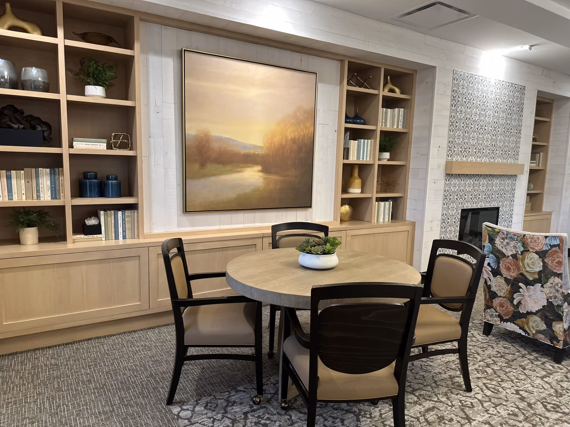 A cozy interior lounge area with a round wooden table surrounded by four chairs with beige cushions. Behind the table is a built-in wooden bookshelf filled with books, decorative items, and plants. A large framed painting of a serene landscape hangs on the wall above the bookshelf. To the right, there is a fireplace with a patterned tile surround and a floral upholstered armchair nearby. The room has a carpeted floor with a subtle pattern.