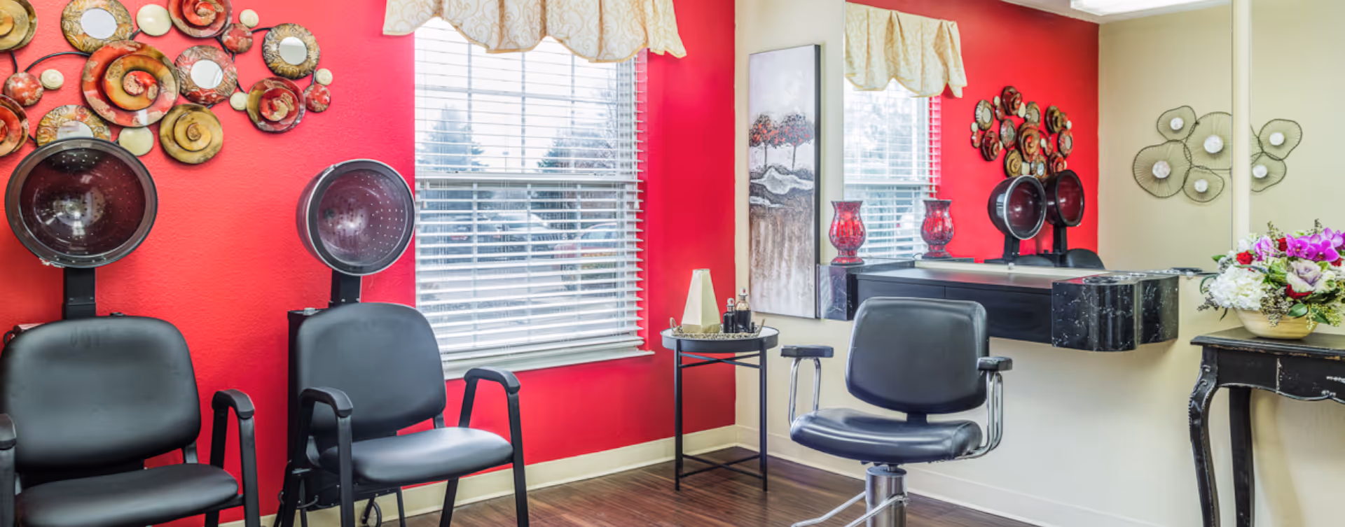 Interior of a salon area with two black hair drying chairs against a red wall, a window with blinds and valance, a black salon chair in front of a large mirror, decorative wall art, and a small table with flowers and vases.