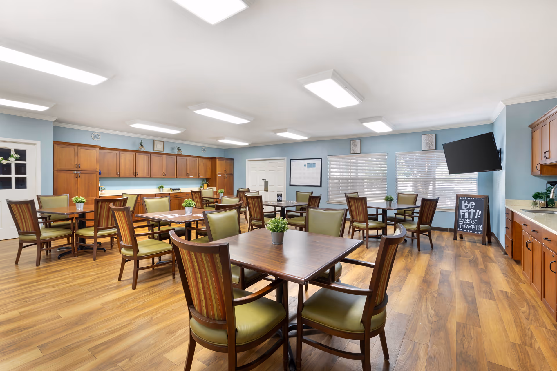 A bright and spacious dining room with multiple wooden tables and chairs with green cushions. The room has light blue walls, wooden flooring, and large windows with blinds. There are cabinets along the walls, a wall-mounted TV, and a chalkboard sign that reads 'Be Fit!! Exercise Today @ 9:30'. Small potted plants are placed on each table.