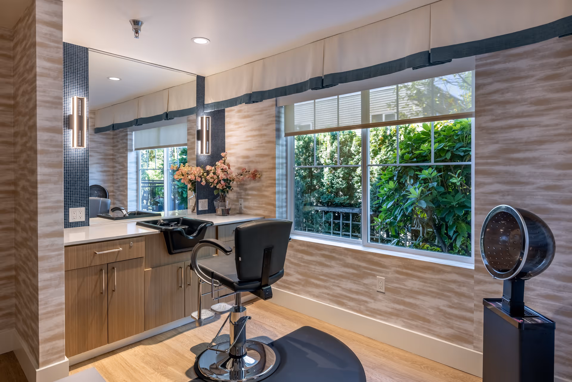 A bright and modern hair salon area with a black salon chair, a hair washing sink, a large mirror with two vertical lights on either side, and a window showing green foliage outside. There is a hair dryer on a stand to the right and a vase with pink flowers on the countertop.