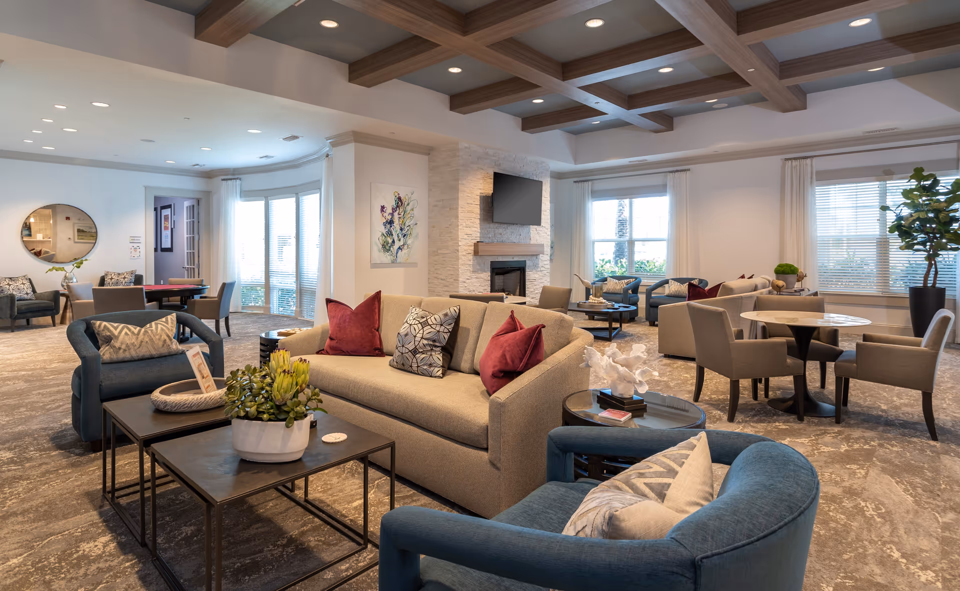 A spacious and well-lit living room area in a senior living facility with comfortable seating including a beige sofa with red and patterned pillows, blue armchairs, and several tables with decorative plants and objects. The room features a coffered ceiling with recessed lighting, large windows with white curtains, a fireplace with a mounted TV above it, and a round mirror on one wall.