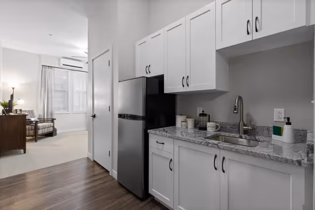 A modern kitchenette with white cabinets, a granite countertop, a stainless steel refrigerator, and a sink. The kitchenette opens into a living area with a wooden chair, a side table with a lamp, and a window with blinds and curtains.