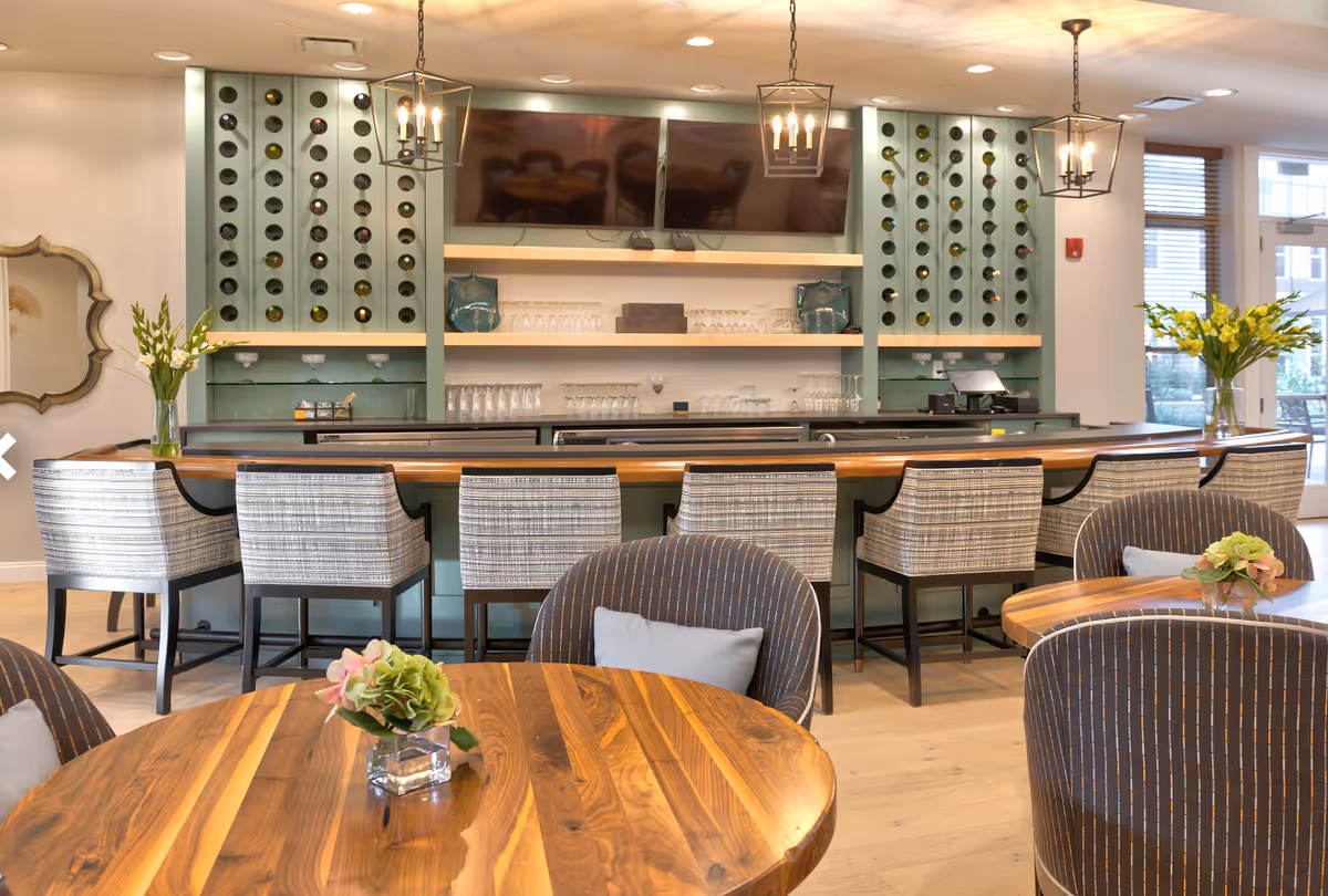 A modern bar area with a wooden countertop and six upholstered bar stools. Behind the bar are shelves with wine bottles, glassware, and two flat-screen TVs mounted on the wall. The room has pendant lights hanging from the ceiling and round wooden tables with cushioned chairs in the foreground. There are vases with flowers on the tables and near the bar, and large windows letting in natural light.