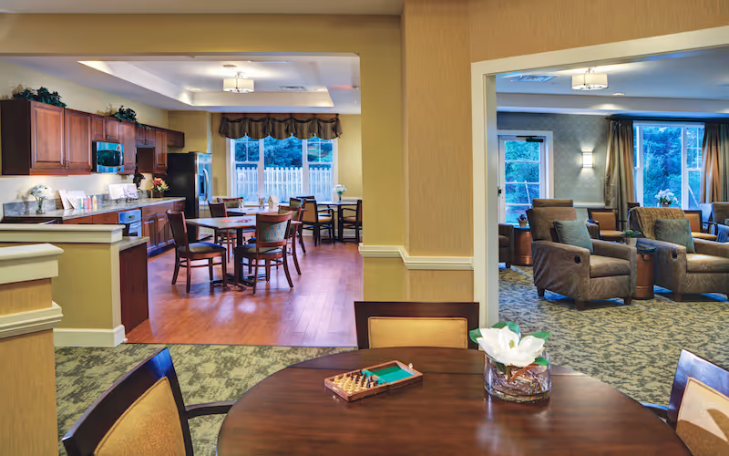 Open communal dining area and adjacent lounge with wooden tables and chairs, a kitchen with cabinetry, and upholstered armchairs.