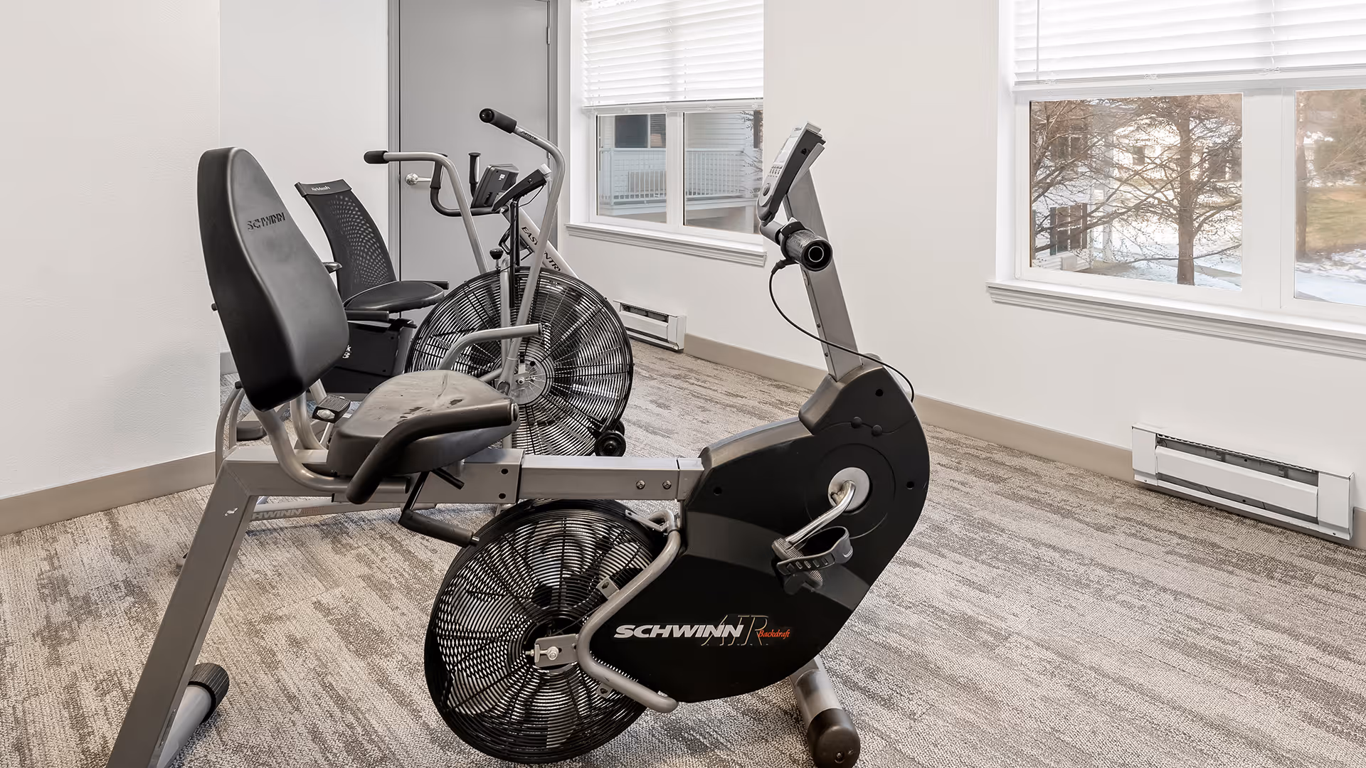 A small exercise room with two stationary recumbent exercise bikes positioned near large windows with white blinds, allowing natural light to fill the space. The room has light-colored walls and carpeted flooring.