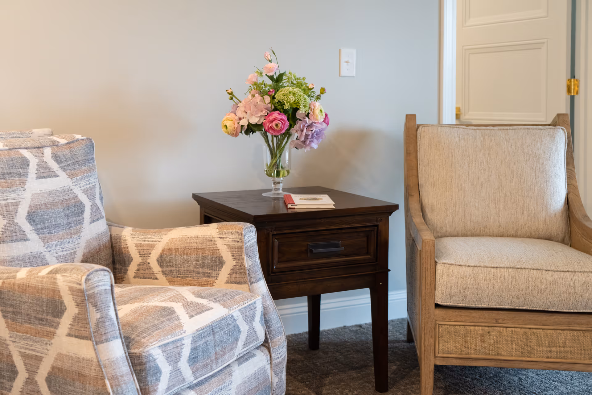 Two armchairs with a wooden side table between them. The side table has a glass vase with a bouquet of colorful flowers and a small book or booklet on top. The background shows a light-colored wall with a power outlet and a partially open door.