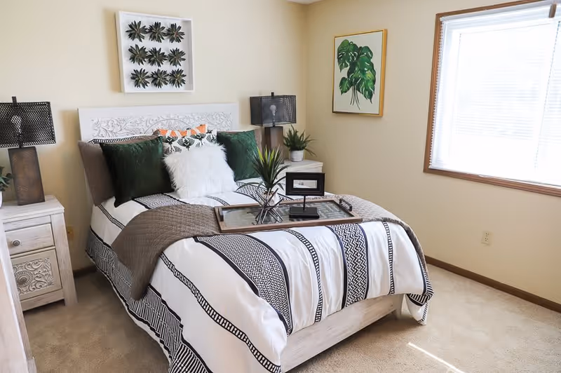 A bright bedroom with a neatly made bed featuring a white and black patterned comforter, green and white pillows, and a brown throw blanket. There are two bedside tables with matching lamps, a framed botanical print on the wall, and a window with blinds letting in natural light.