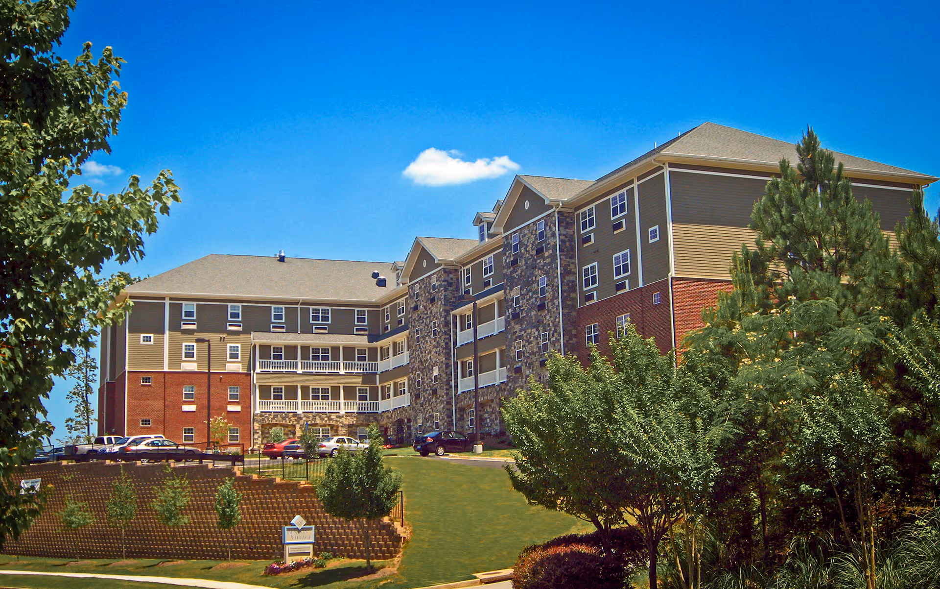 Exterior view of a multi-story senior living facility building with a combination of brick, stone, and siding. The building is surrounded by green trees, shrubs, and a well-maintained lawn under a clear blue sky.