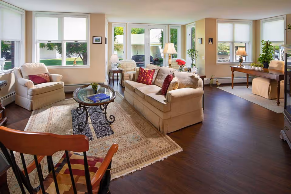 A bright and cozy living room in a retirement community featuring a beige sofa with red and patterned cushions, a matching armchair, a glass coffee table on an ornate rug, and a wooden chair in the foreground. Large windows and glass doors let in natural light and offer a view of greenery outside. There is a wooden desk with a chair and plants near the windows, and lamps providing warm lighting throughout the room.