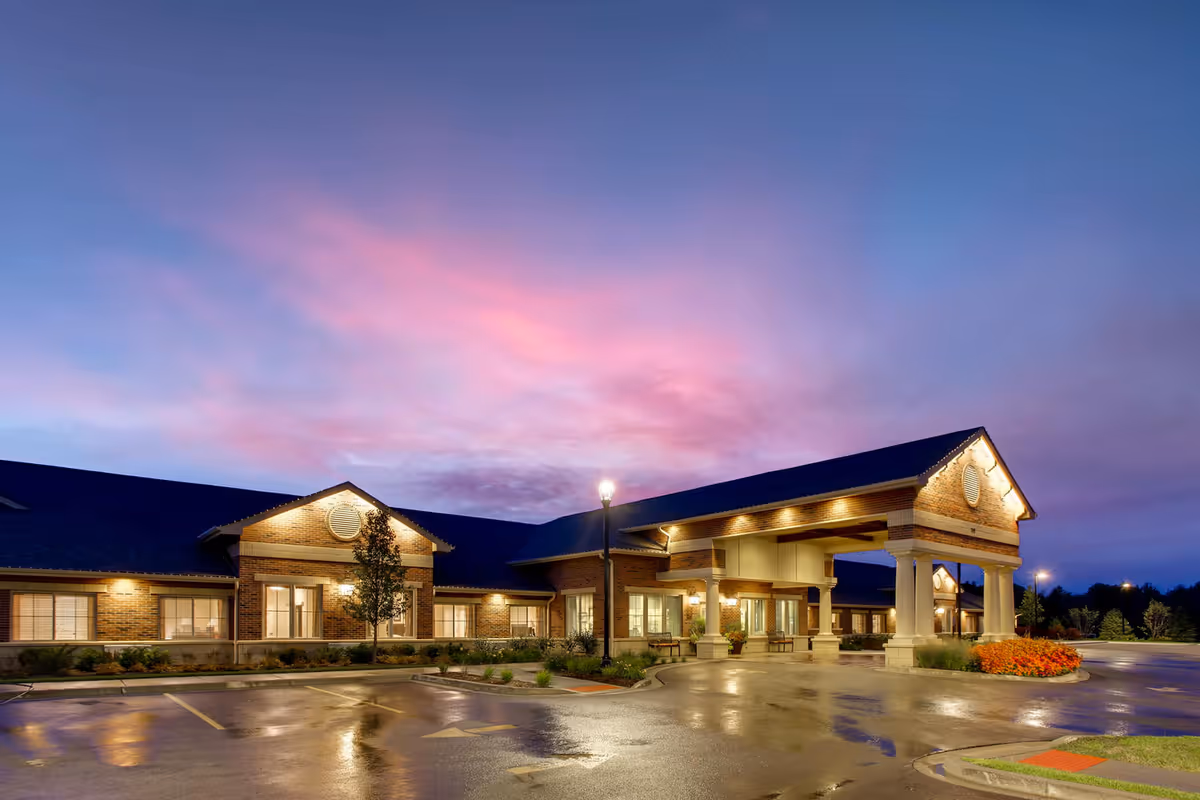 Exterior view of Maple Glen Memory Care Community building at dusk with illuminated entrance, brick facade, and a wet parking lot reflecting the lights under a colorful purple and pink sky.