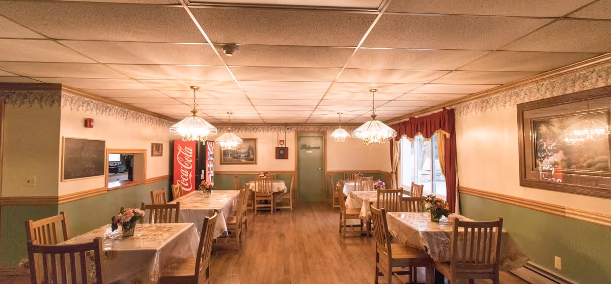 A dining room with several wooden tables and chairs arranged neatly. Each table is covered with a floral tablecloth and has a small flower arrangement. The room has a wooden floor, beige and green walls with a decorative border near the ceiling, and hanging light fixtures. A Coca-Cola vending machine is visible in the back corner near a door labeled 'Restrooms'. Large windows with red curtains allow natural light into the room.
