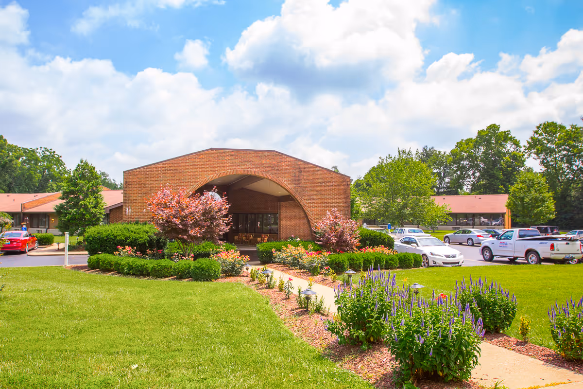 Front entrance of a brick senior living facility with landscaped gardens, a walkway, and parked cars.