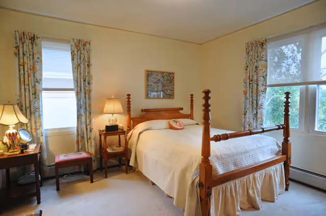 Sunlit bedroom with a wooden four-poster bed, bedside tables and lamps, floral curtains, and a small dressing table.