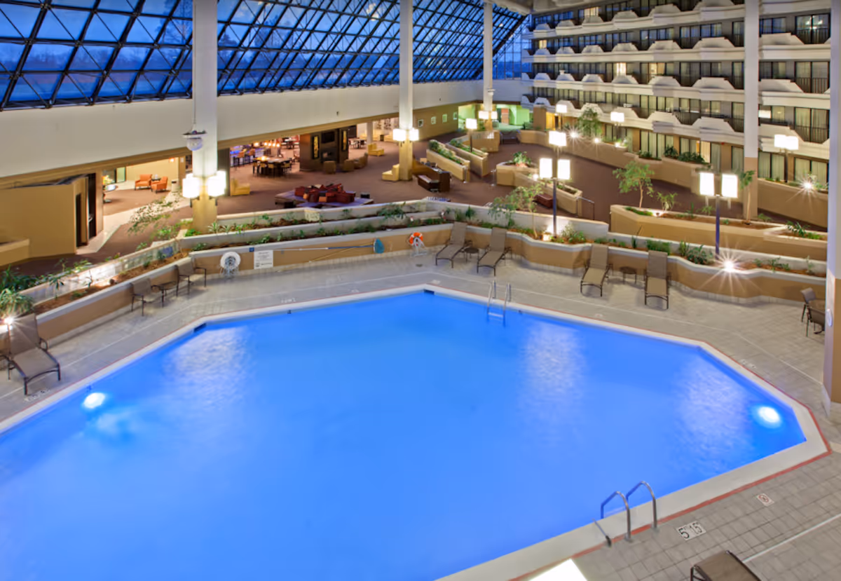 Indoor swimming pool with blue water surrounded by lounge chairs and plants, located inside a large atrium with a glass ceiling and multiple floors of balconies overlooking the pool area.