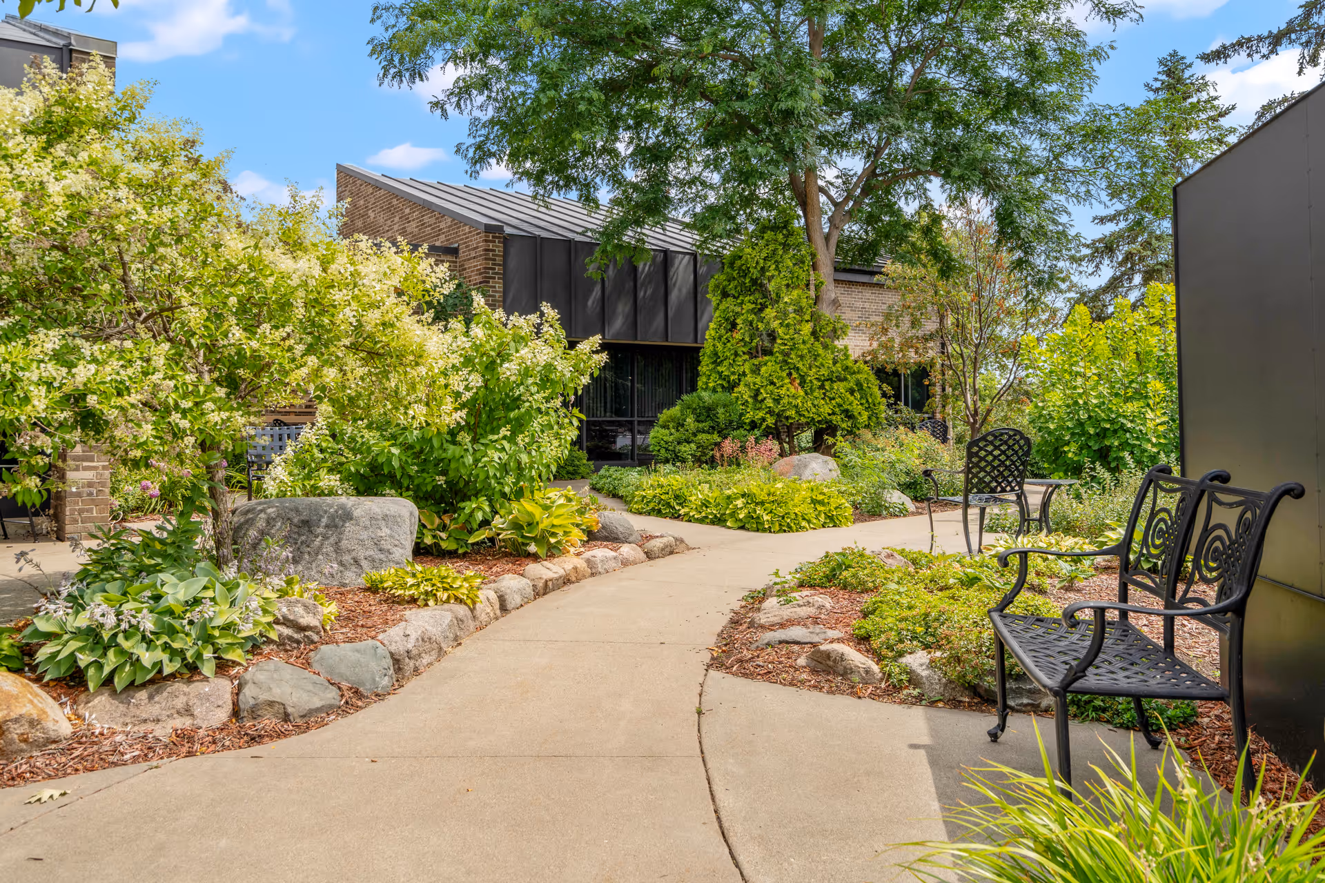 A landscaped outdoor courtyard with a paved walkway, garden beds, and metal benches in front of a brick building.