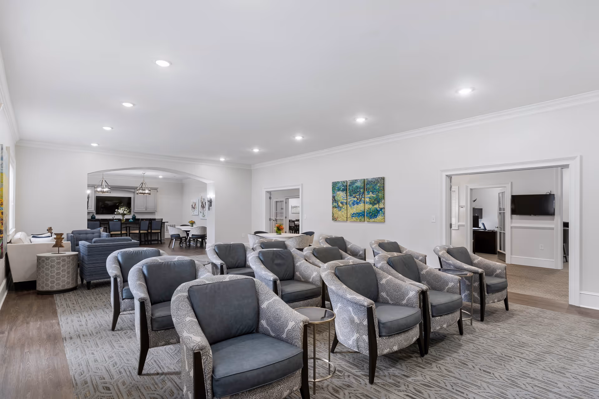 A spacious and well-lit common area with multiple upholstered armchairs arranged in rows facing forward. The room has light-colored walls, recessed ceiling lights, and a patterned carpet. In the background, there is a dining area with tables and chairs, and a kitchen area with a counter and bar stools. A colorful triptych painting hangs on the wall, and an adjacent room with a wall-mounted TV is visible through an open doorway.