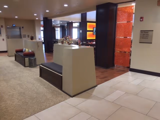 Interior view of a senior living facility hallway with seating areas, decorative plants, and a sign indicating room numbers on the first floor. The space features a mix of carpet and tile flooring, with warm lighting and modern decor.