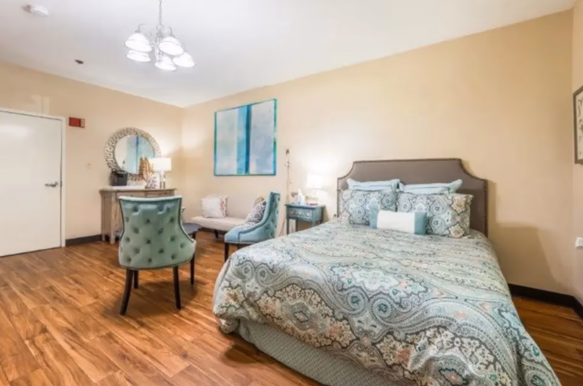 A cozy bedroom with a large bed covered in a patterned blue and beige bedspread, two matching pillows, and a bolster pillow. The room has wooden flooring, beige walls, and soft lighting from a bedside lamp. There is a small blue nightstand next to the bed, a green upholstered chair, a small sofa with cushions, a wooden dresser with a round decorative mirror above it, and a ceiling light fixture with multiple bulbs.