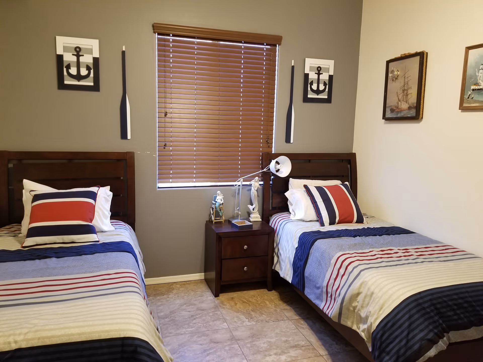 Bedroom with two twin beds featuring nautical-themed bedding and wall decor, a nightstand with a lamp between them and a window with closed blinds.