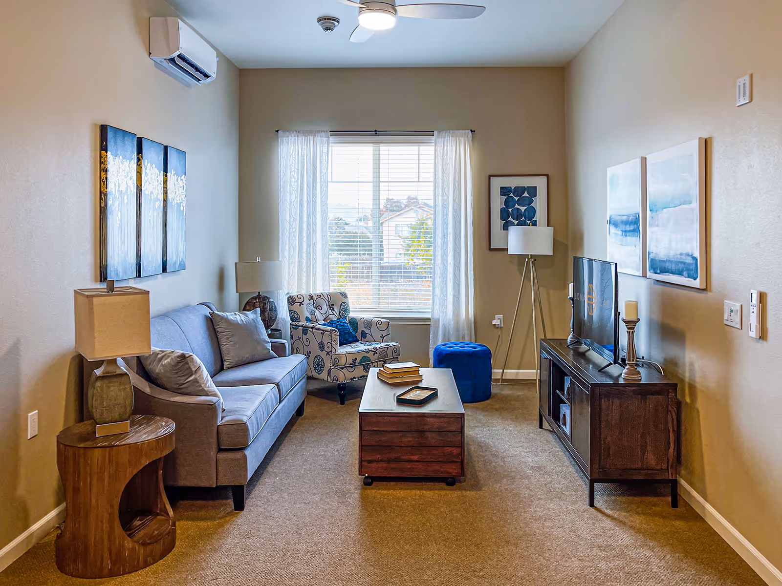 Bright furnished living room with a sofa, patterned armchair, coffee table, TV console, and a window with curtains.
