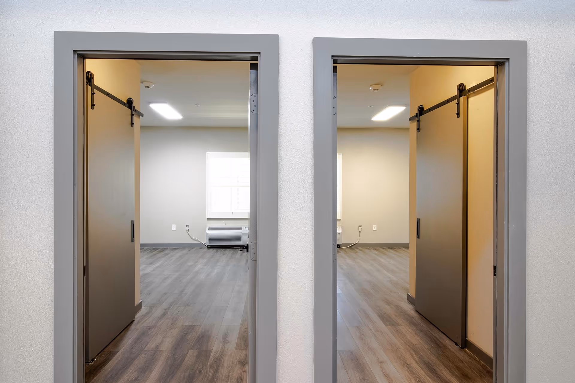 View through two adjacent doorways with sliding barn-style doors partially open, showing empty rooms with wood flooring, beige walls, a window with blinds, and a wall-mounted air conditioning unit.