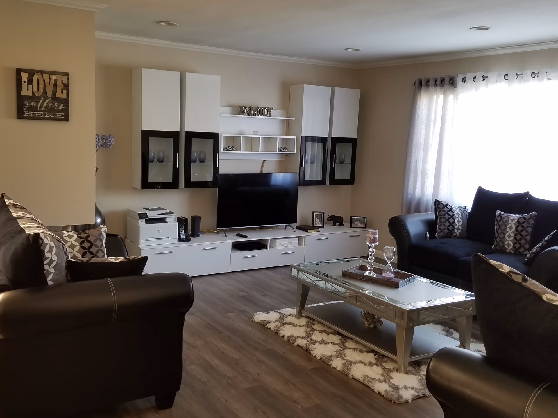 A cozy living room with dark leather sofas adorned with patterned cushions, a glass coffee table on a patterned rug, a flat-screen TV mounted on a white entertainment unit with shelves and cabinets, and a window with light curtains allowing natural light to fill the room. A wall decoration reads 'LOVE gathers HERE'.