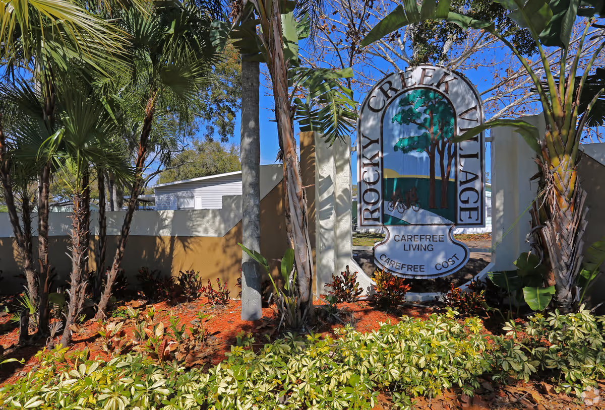 Decorative entrance sign for Rocky Creek Village surrounded by palm trees and landscaped plants under a blue sky.