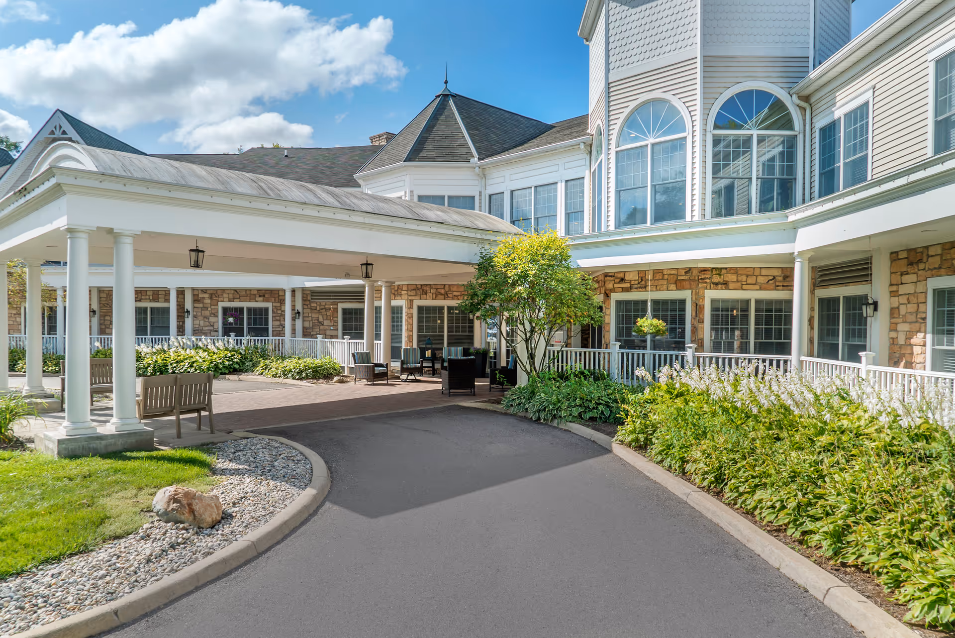 Front entrance of a senior living building with a covered porte-cochere, benches, and landscaped beds.