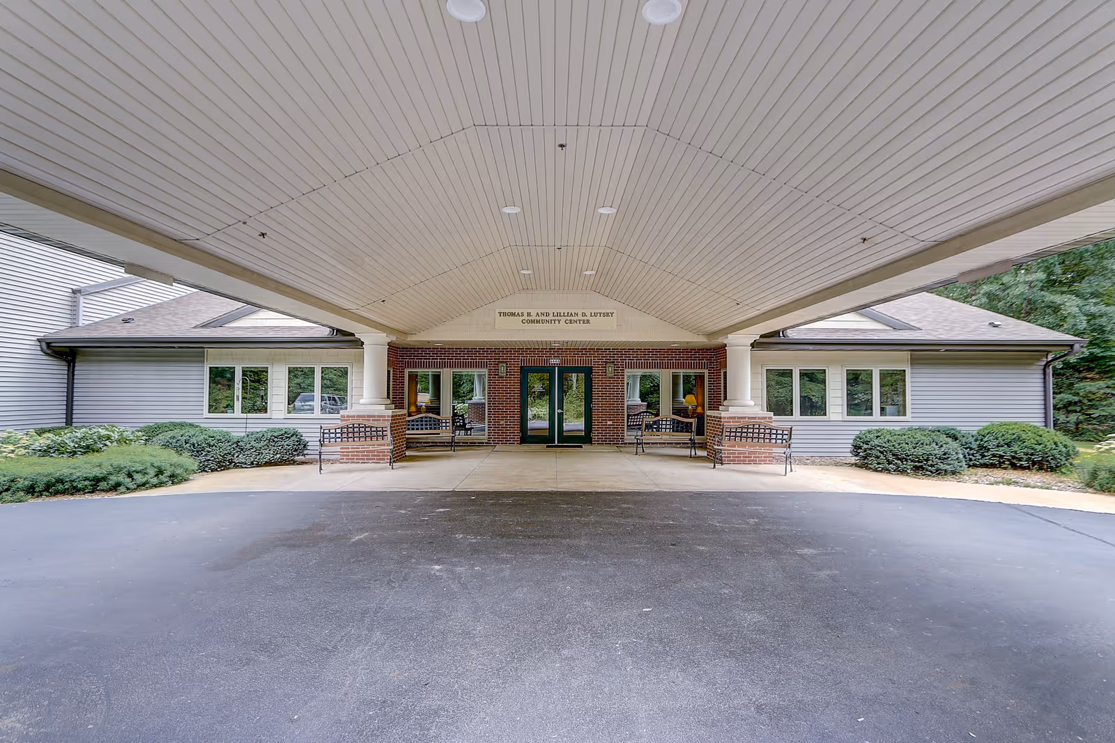 Entrance to the Thomas H. and Lillian B. Lutsey Community Center at MasonWoods, featuring a covered driveway with white ceiling panels, brick pillars, benches on either side, and glass double doors flanked by windows. Surrounding the entrance are bushes and trees.