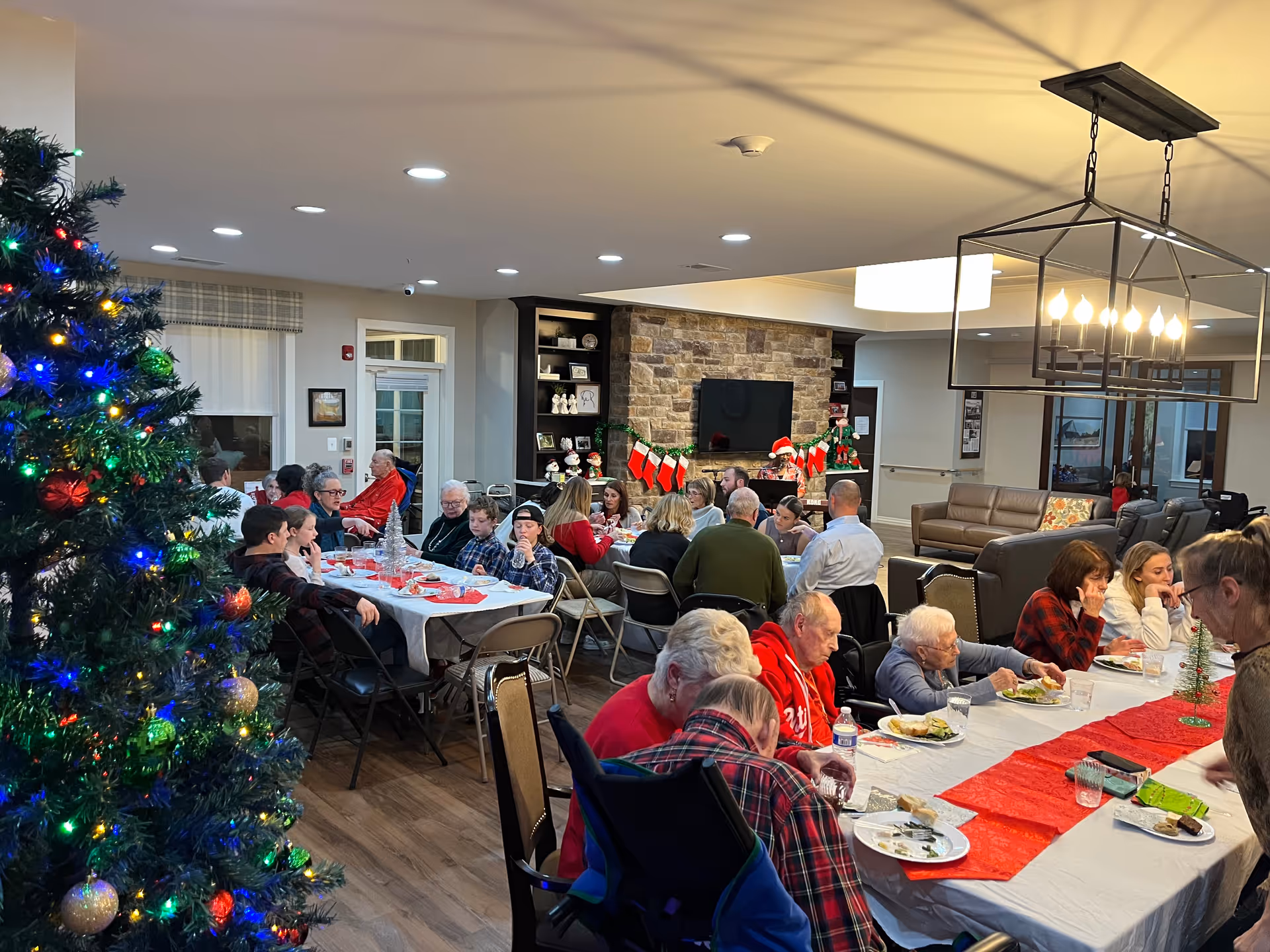 Seniors and family members gathered around long tables in a decorated common room enjoying a holiday meal with a Christmas tree nearby.
