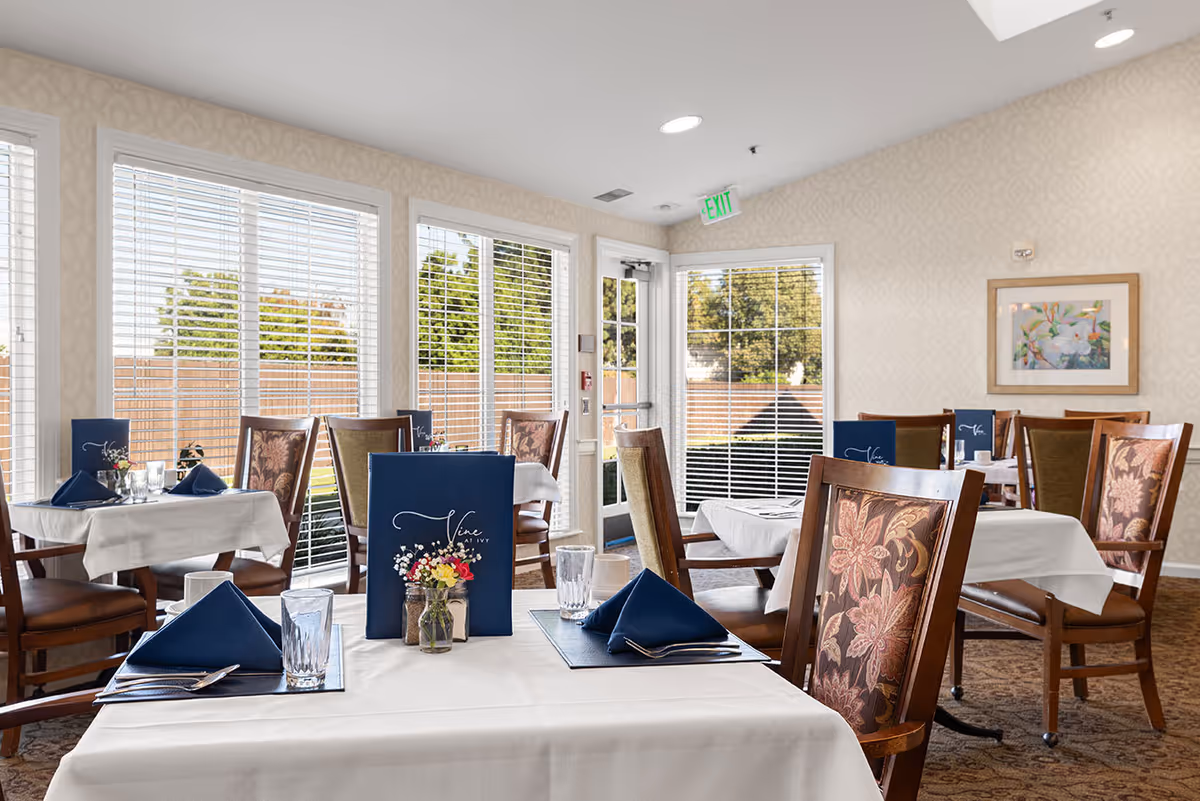 A bright dining room with several tables covered in white tablecloths, each set with blue napkins, glasses, and menus. The chairs have floral patterned upholstery. Large windows with white blinds allow natural light to fill the room, and a framed floral painting hangs on the wall.