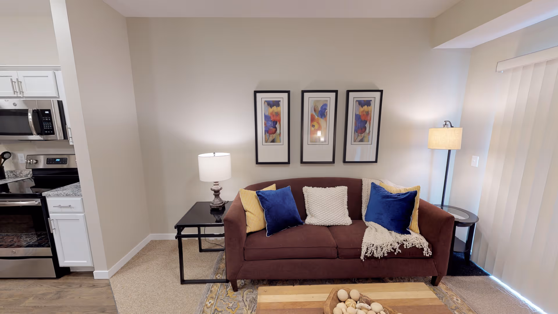 A cozy living room area featuring a brown sofa with blue, yellow, and white pillows, a white throw blanket, two side tables with lamps, three framed abstract paintings on the wall, and a view of a kitchen area with stainless steel appliances and white cabinets.