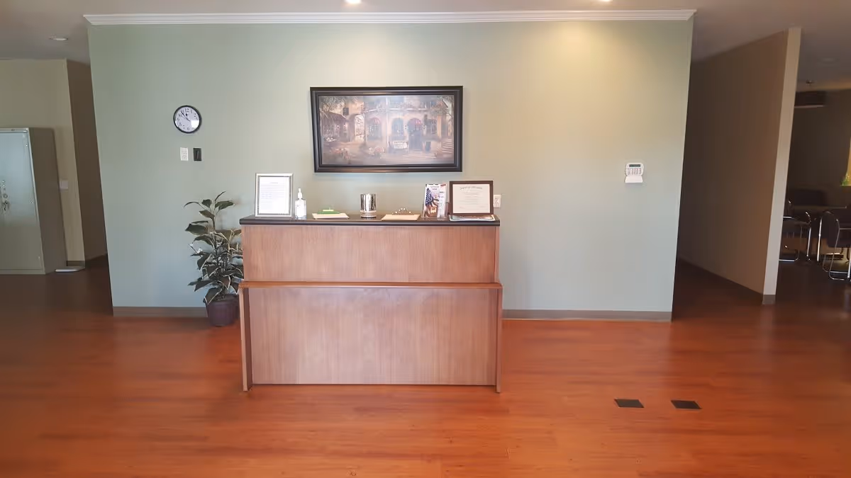 Reception area with a wooden desk in the center, a potted plant to the left, a clock on the wall, and a framed picture hanging above the desk. The floor is wooden and the walls are painted light green.