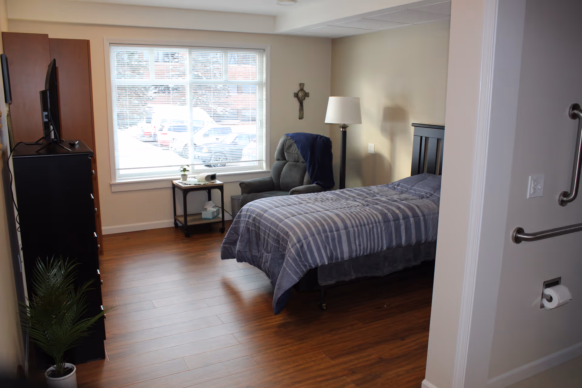 A tidy bedroom with a single bed, recliner chair, side table, TV cabinet and large window overlooking a parking lot.
