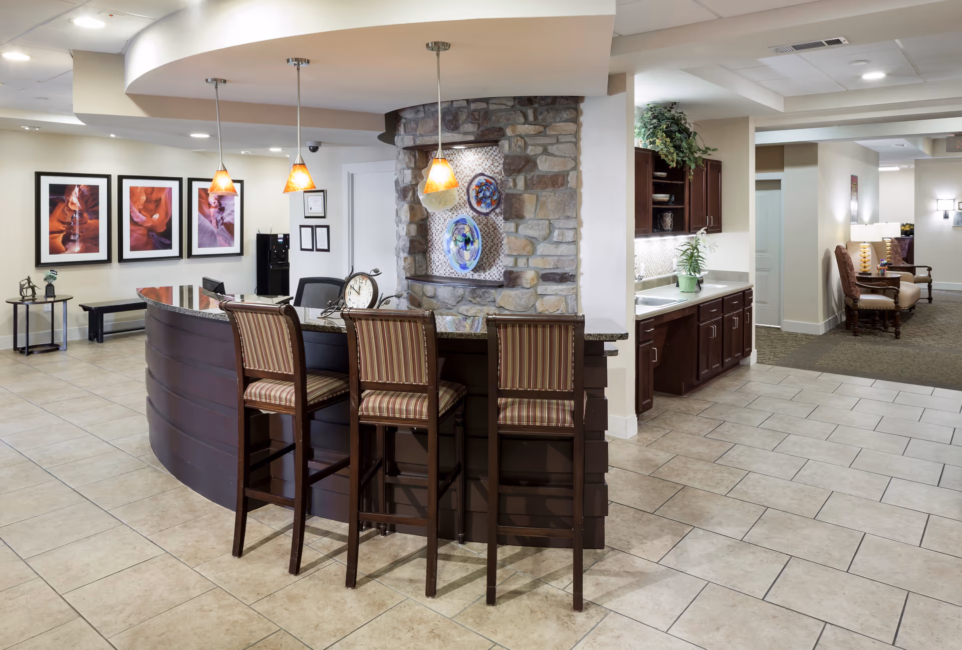 Spacious senior living communal interior featuring a curved reception/bar counter with three high stools, pendant lights, a kitchenette, and seating areas.