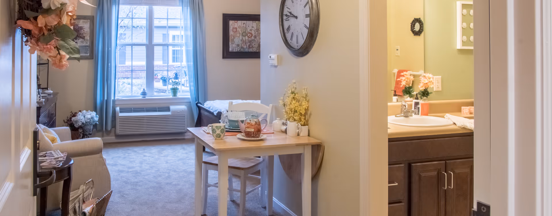 A cozy living area with a small wooden table set for tea, a white chair, a large wall clock, and a window with blue curtains letting in natural light. To the right, there is a partial view of a bathroom sink with a mirror and countertop. The room has soft beige walls and carpeted flooring.