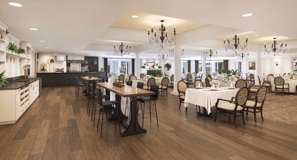 A spacious and elegant dining room with wooden floors, multiple tables covered with white tablecloths, and chairs with patterned upholstery. The room features chandeliers hanging from the ceiling, large windows allowing natural light, and a kitchen area with a counter and bar stools on the left side.