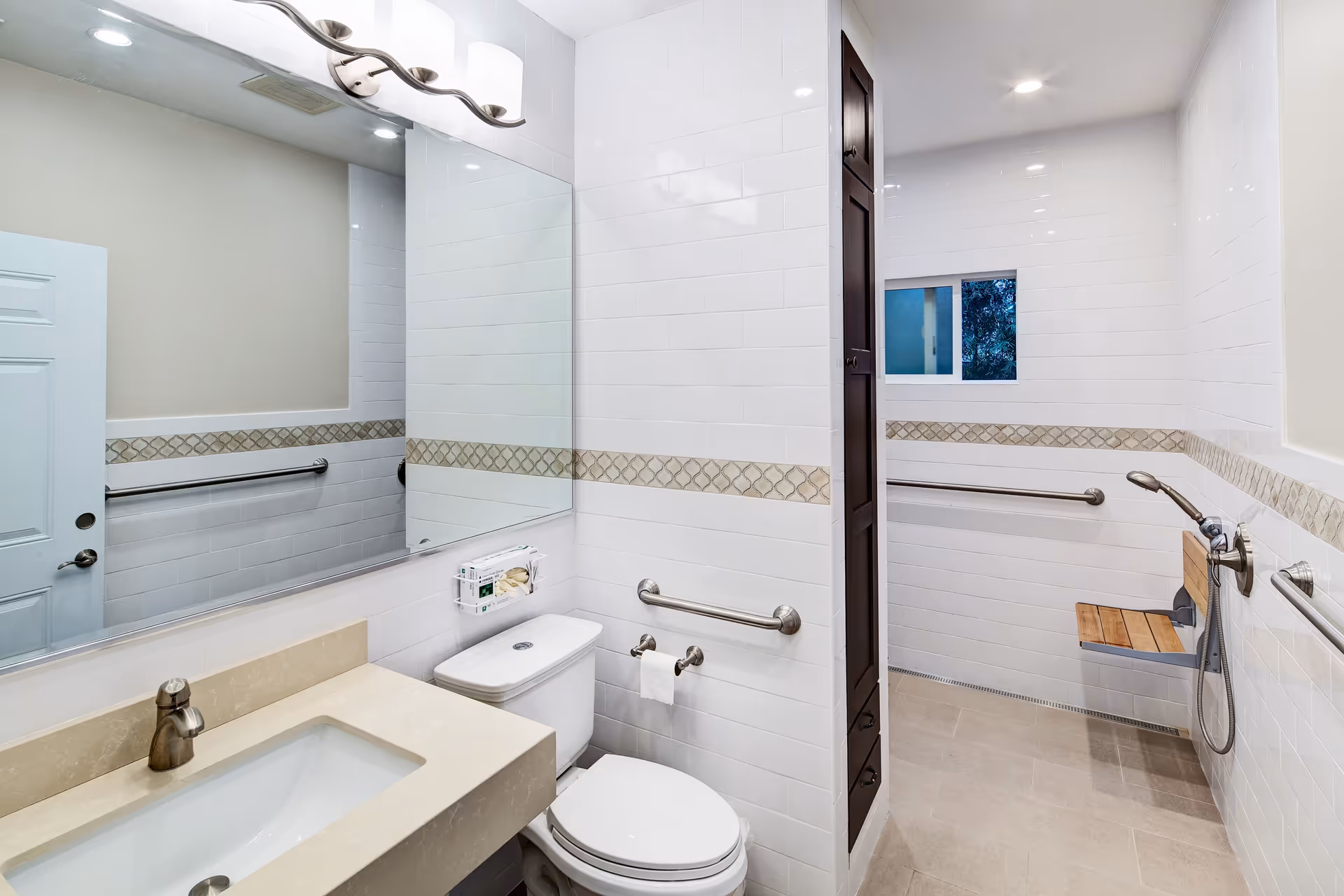 A modern bathroom with white tiled walls and beige floor tiles. The bathroom features a beige countertop with an integrated sink, a large mirror above the sink, a toilet with a toilet paper holder and grab bars on the wall. There is a walk-in shower area with a wooden fold-down seat, a handheld showerhead, grab bars, and a small window providing natural light.