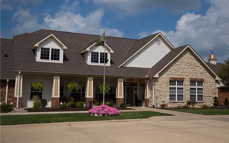 Exterior view of a single-story senior living facility building with a combination of brick, stone, and siding. The building has a dark shingled roof with dormer windows, a flagpole with a green flag, and a landscaped front area featuring pink flowers and green shrubs under a partly cloudy sky.