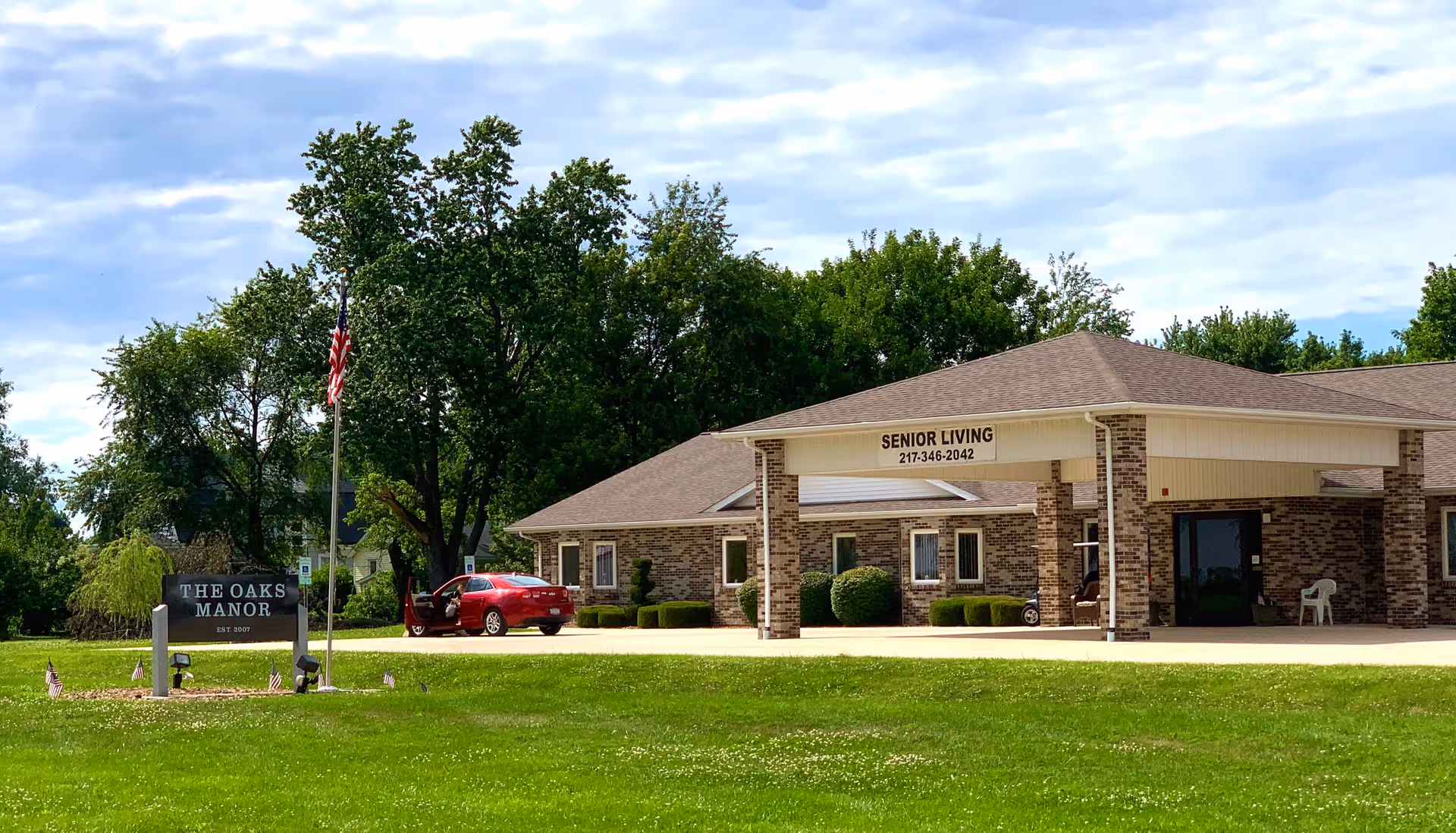 Front exterior of The Oaks Manor senior living facility with a sign, flagpole, parked car, and covered entrance.