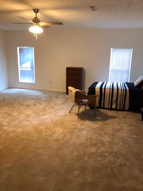 A sparsely furnished bedroom with beige carpet, two narrow windows with blinds, a ceiling fan with light, a wooden dresser, a single bed with a black and white striped blanket, and a wicker chair with a cushion.