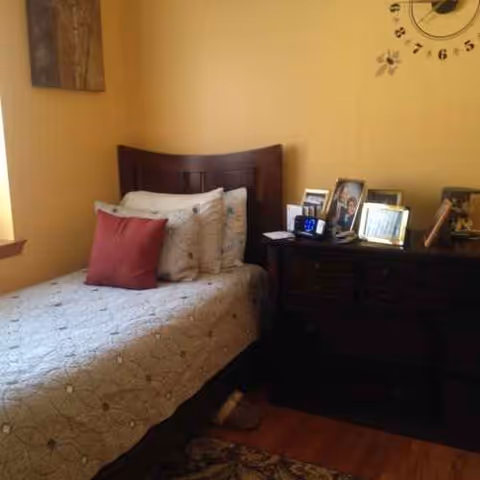 Bedroom in a senior living facility with a bed, pillows, and a dresser with photos