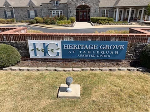 A blue sign reading 'HERITAGE GROVE AT TAHLEQUAH ASSISTED LIVING' in front of a stone building entrance and landscaped lawn.