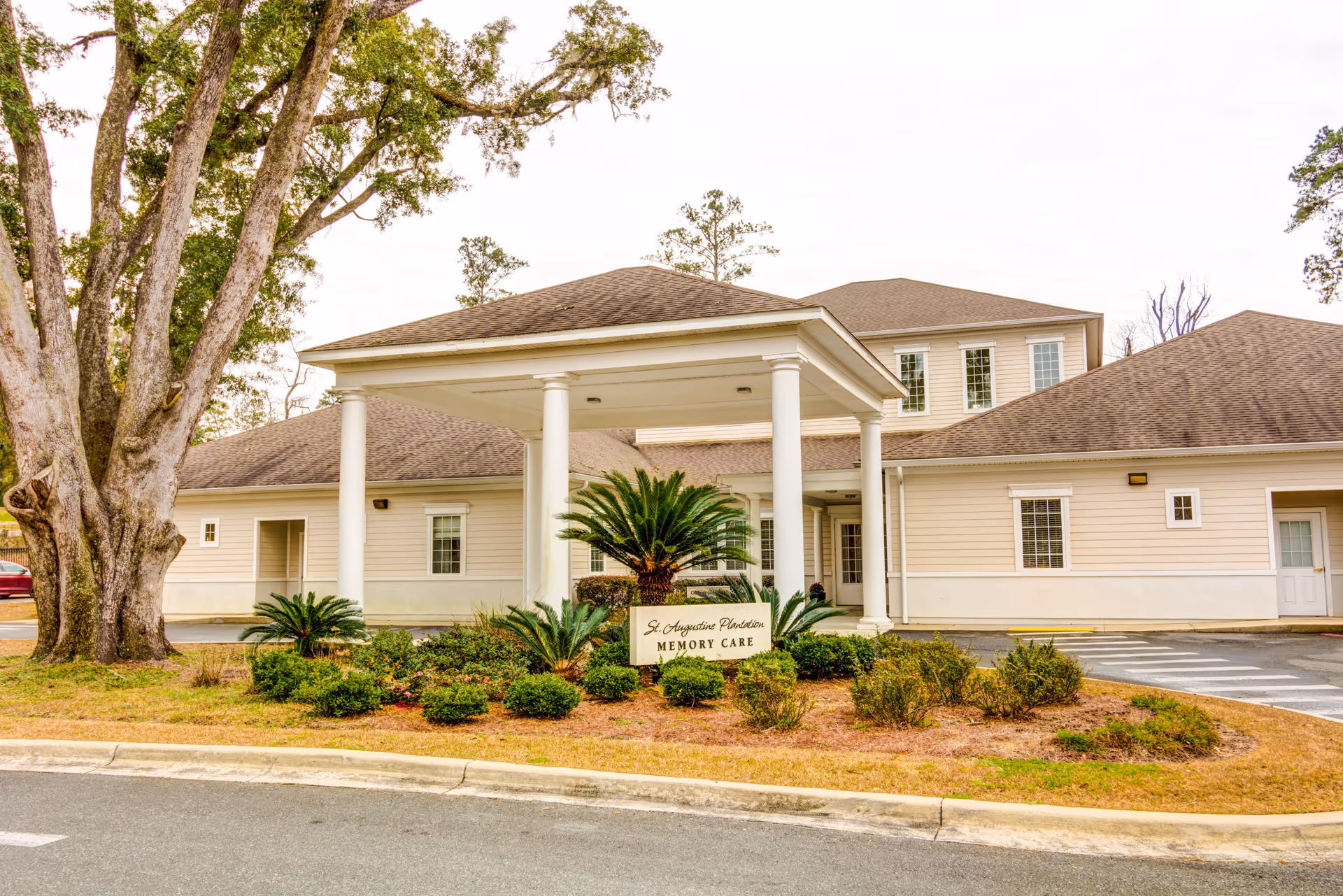 Exterior view of St. Augustine Plantation Memory Care building with beige siding, a covered entrance supported by white columns, landscaping with bushes and palm plants, and large trees nearby.