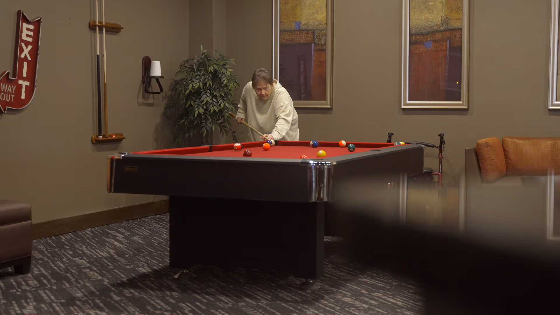 An elderly person playing pool on a red felt pool table in a cozy room with brown walls, a potted plant, framed artwork, and a brown couch. There is a vintage red exit sign on the wall.