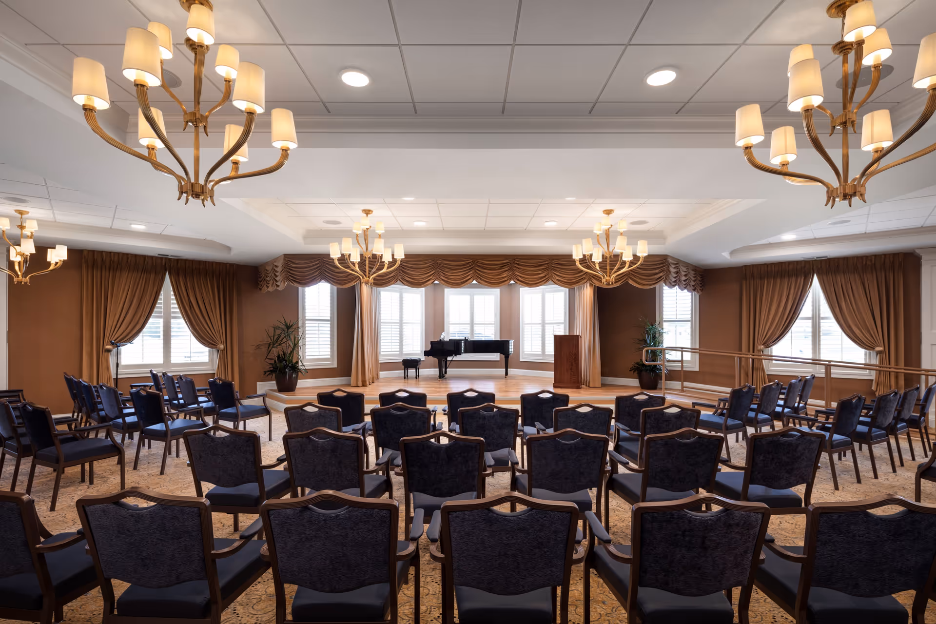 A large, elegant room set up for a presentation or performance with rows of chairs facing a small stage. The stage has a grand piano and a wooden podium. The room features large windows with brown curtains, beige walls, and multiple chandeliers hanging from the ceiling.