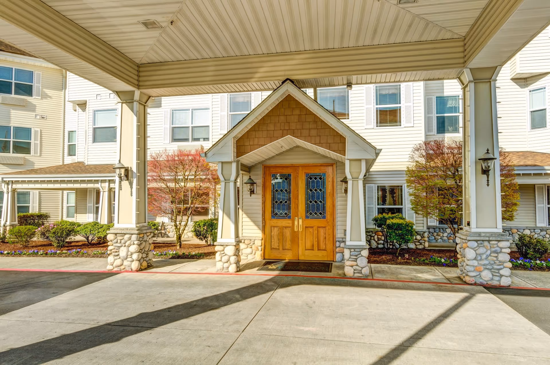 Entrance of a senior living facility with a covered driveway supported by columns with stone bases, wooden double doors with decorative glass panels, and landscaping including bushes and small trees.