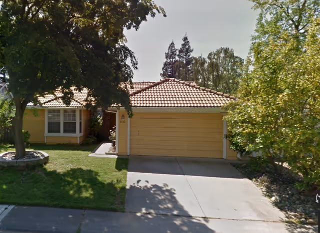 Front exterior of a single-story house with a tan two-car garage and surrounding trees.
