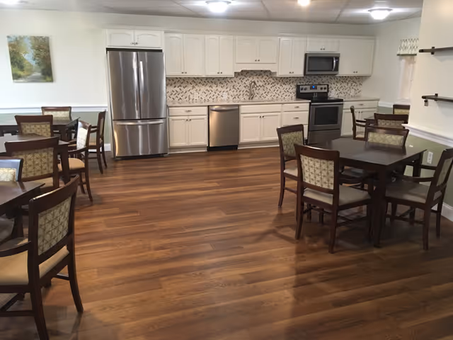 A spacious kitchen and dining area with wooden floors, white cabinets, stainless steel refrigerator, dishwasher, oven, and microwave. Several wooden dining tables with cushioned chairs are arranged around the room. A painting hangs on the wall to the left and a window with a valance is on the right.